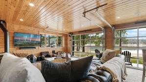 A spacious room with a wooden ceiling, large windows, a black leather sofa, and a mounted TV. - The Black Cormorant | On the Lake (Big Bear Lake)