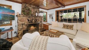 A cozy living room with a stone fireplace, a large window, and a comfortable sofa. - The Black Cormorant | On the Lake (Big Bear Lake)