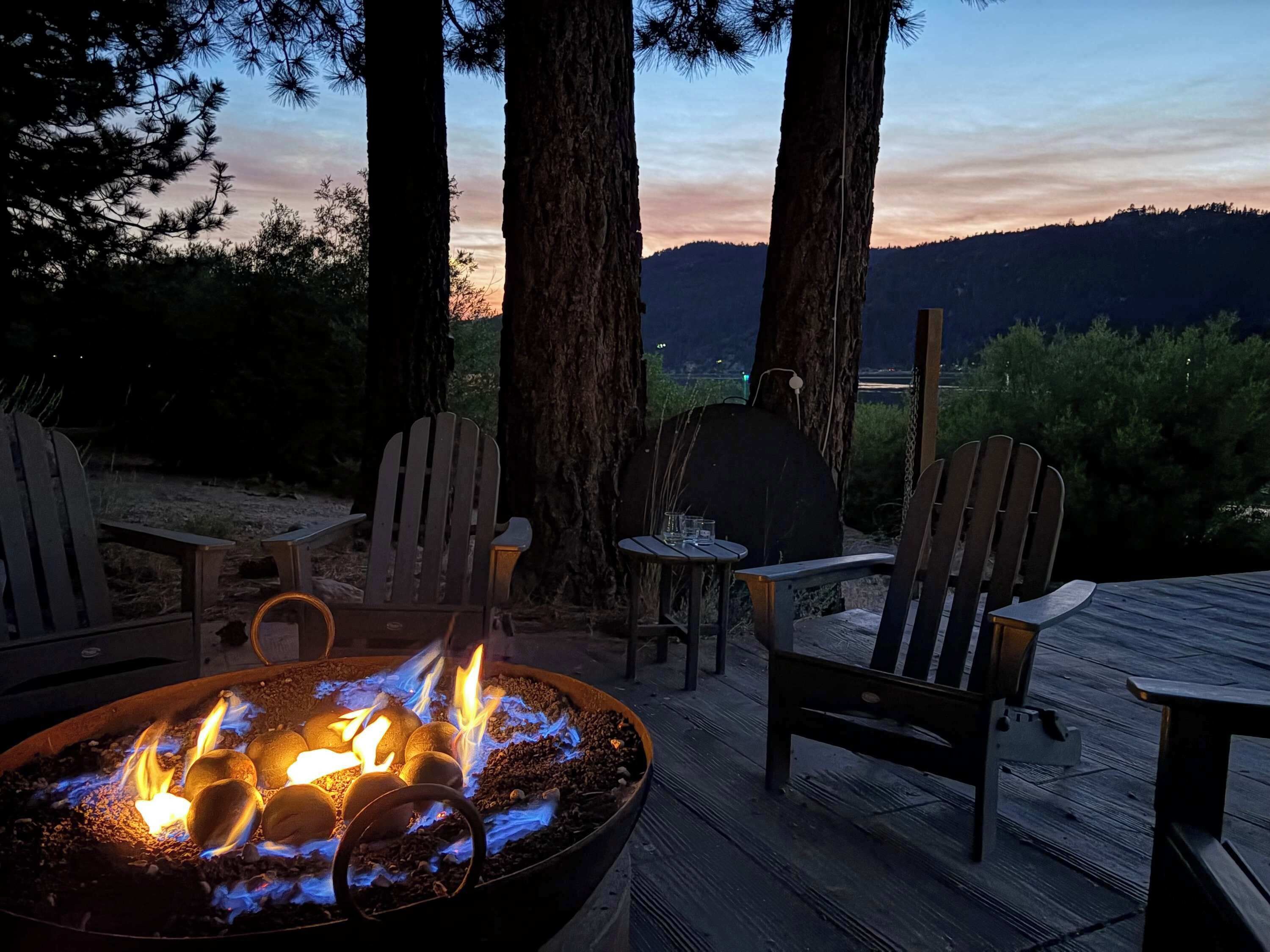 A fire pit with logs, surrounded by wooden chairs and tables, set against a backdrop of trees and a sunset.