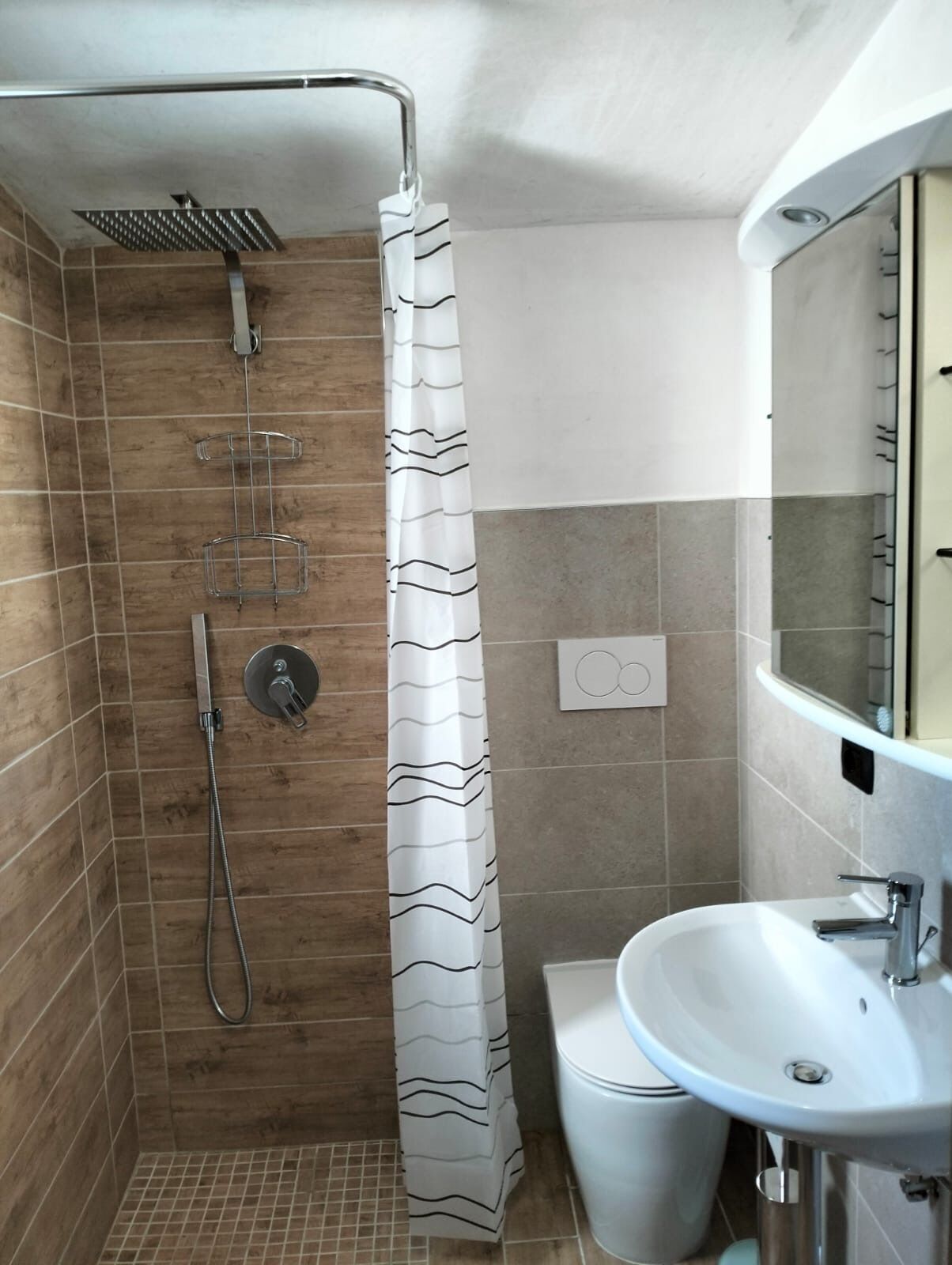Ducha con cabezal de ducha curvo, cabezal de ducha manual y control de pared. Hay una cortina de ducha blanca con un diseño ondulado. Se ve un lavamanos blanco con un espejo encima.