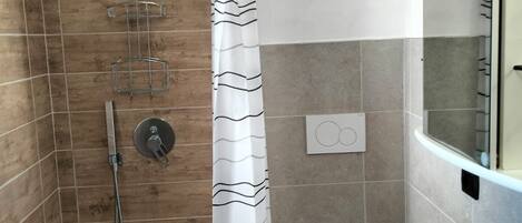 Ducha con cabezal de ducha curvo, cabezal de ducha manual y control de pared. Hay una cortina de ducha blanca con un diseño ondulado. Se ve un lavamanos blanco con un espejo encima.