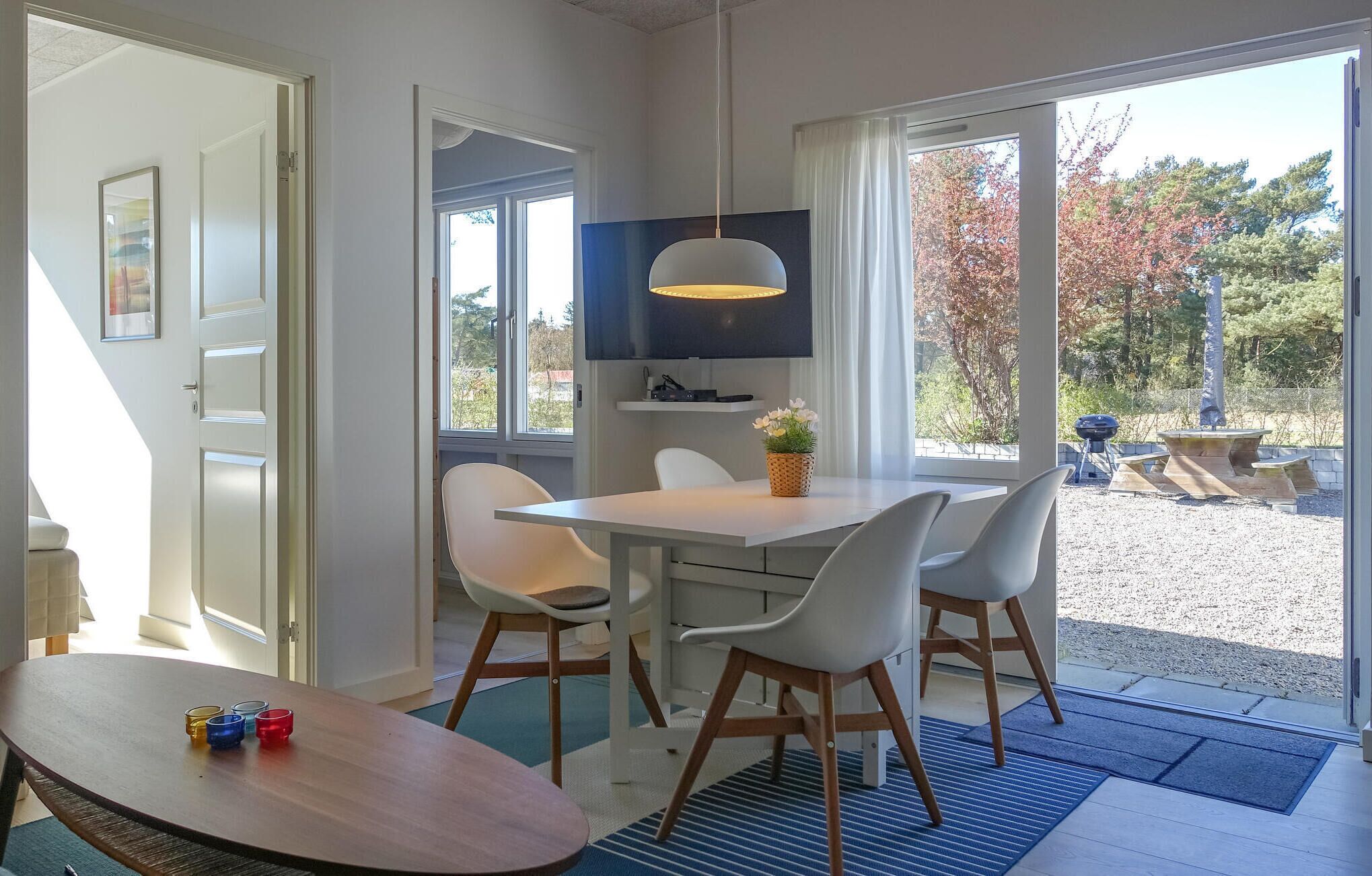 A modern dining area with a white table and chairs, a hanging light fixture, and a view of the outdoors through large windows.