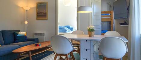 A compact hotel room with a sofa, a dining table with chairs, a small TV, and a bunk bed.
