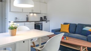 Moderni keittiö, jossa on valkoiset kaapit, ruokailutila valkoisella pöydällä ja tuoleilla sekä sininen sohva värikkäillä koristetyynyillä.