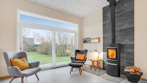 A modern living room with a large sliding glass door leading to a garden, a black wood-burning stove, two armchairs, and a small wooden table with a lamp. - 3 bedroom cozy home in Tarm (Tarm)