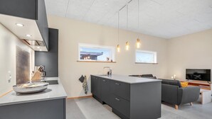 A modern kitchen with a central island, dark cabinetry, and pendant lighting. There is a dining area with a sofa and a flat-screen TV. - 3 bedroom cozy home in Tarm (Tarm)