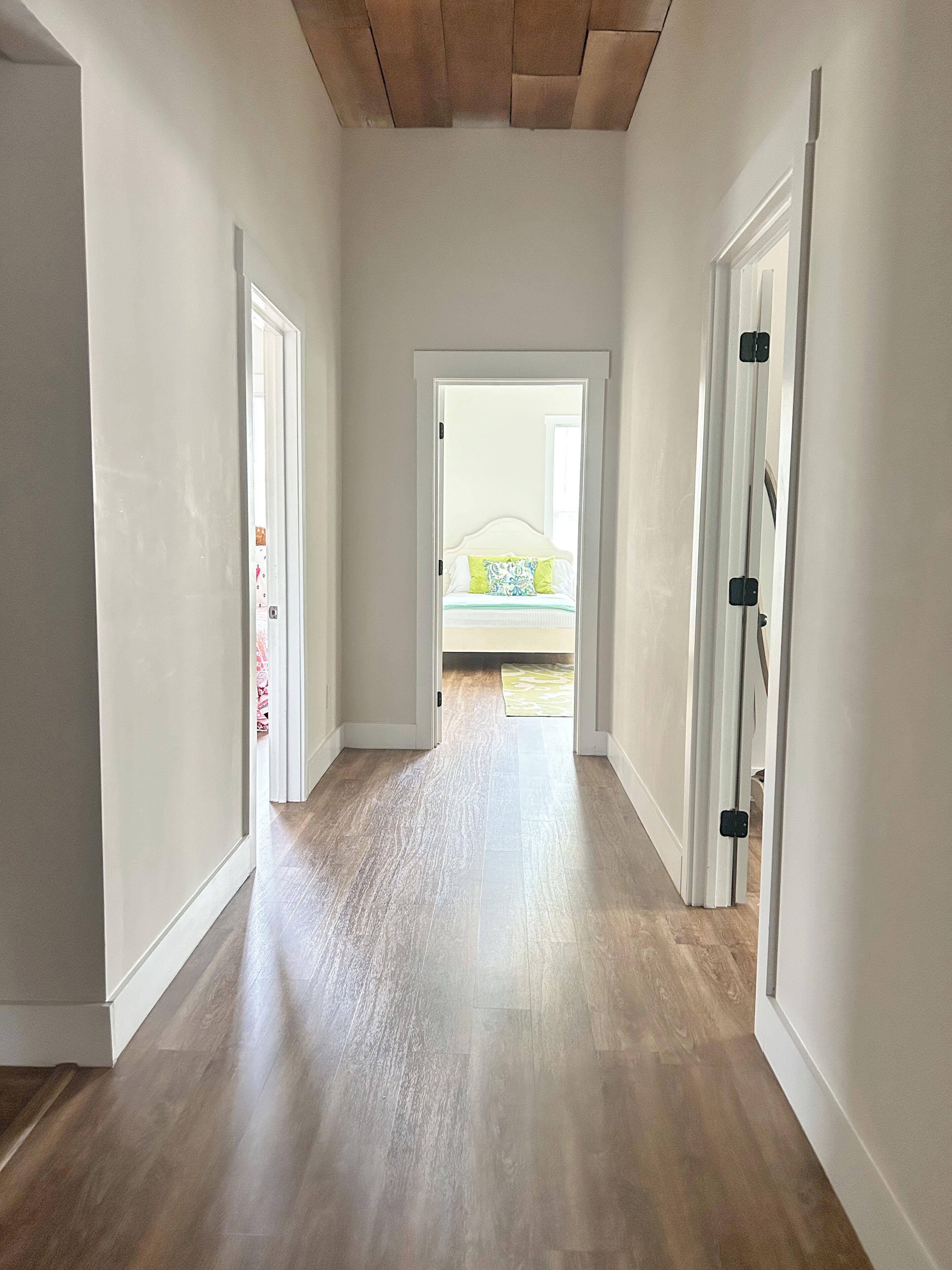 A hallway with wooden flooring and white walls, leading to a bedroom with a bed and a window.
