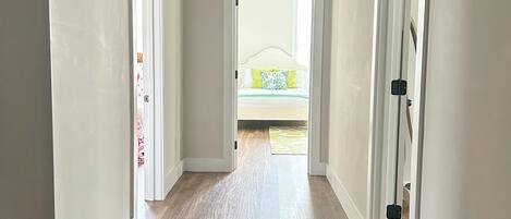 A hallway with wooden flooring and white walls, leading to a bedroom with a bed and a window.