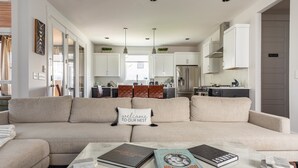 A modern living room with a beige sectional sofa, a coffee table with books, and a kitchen in the background.