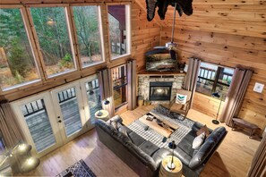 A cozy cabin interior with a stone fireplace, a black leather sofa, and large windows offering a view of the forest. - Mountain Views_Firepit_Hot tub_ BEST location: Between Pigeon Forge & Gatlinburg (Sevierville)