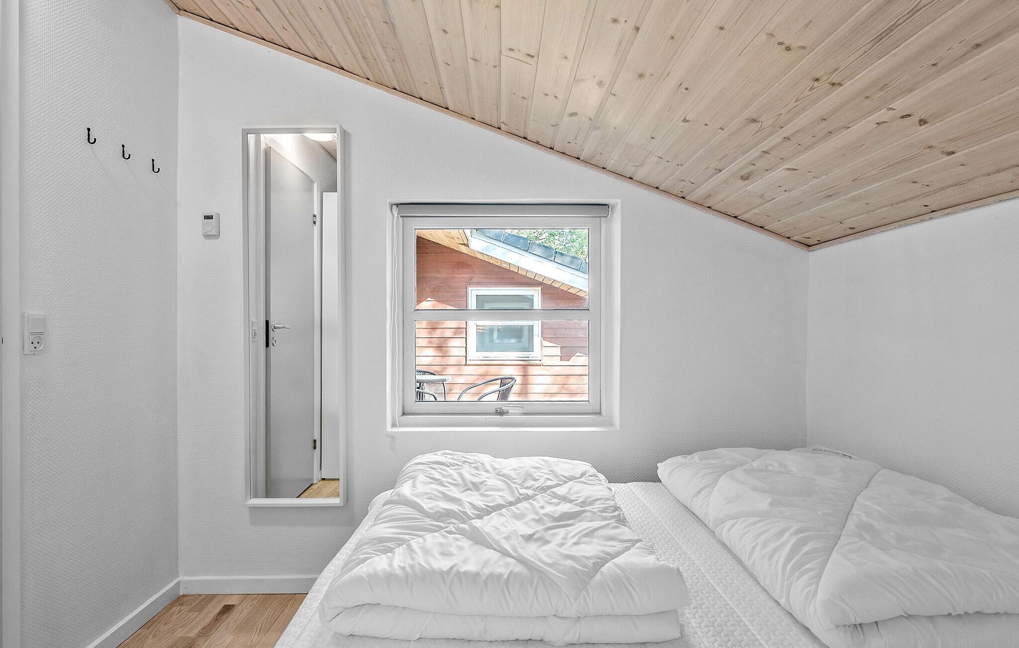 Une chambre avec un plafond en pente, une fenêtre donnant sur une terrasse en bois, un lit avec une literie blanche et une porte dotée d’un miroir.
