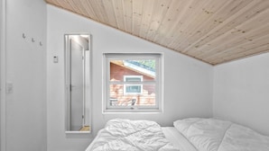 Une chambre avec un plafond en pente, une fenêtre donnant sur une terrasse en bois, un lit avec une literie blanche et une porte dotée d’un miroir.