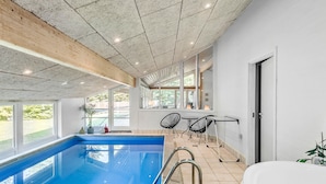 Une piscine intérieure moderne avec des parois en verre transparent, un plafond en bois et un patio ensoleillé doté de sièges extérieurs.