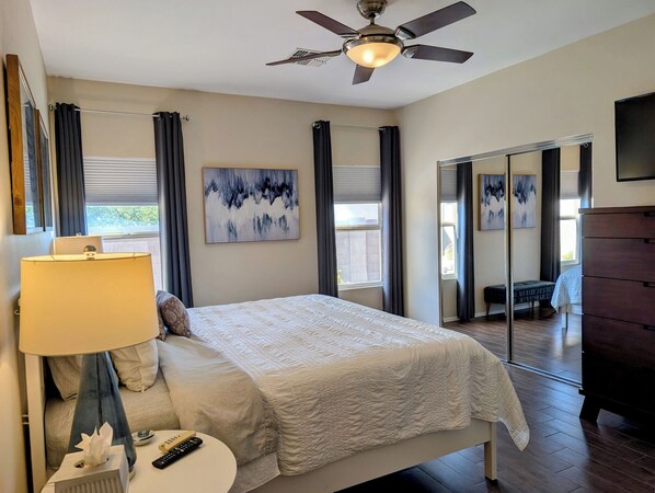 A bedroom with a bed, a bedside table with a lamp, a ceiling fan, and artwork on the walls.