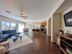 A modern living room with a sofa, coffee table, and TV stand. There is a kitchen area with a dining table and chairs, and a ceiling fan.