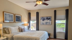 Ein Schlafzimmer mit Bett, Nachttischen, Deckenventilator, Kunstwerken und einem Fenster mit Vorhängen.