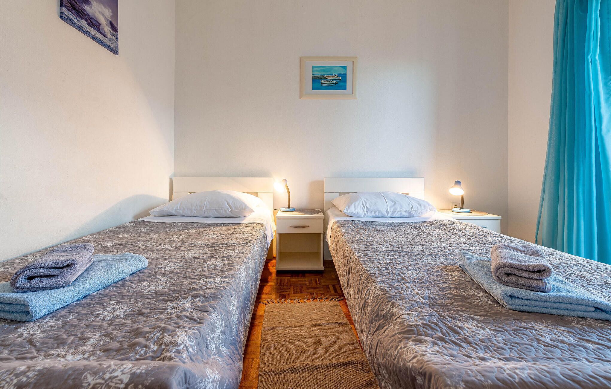 A hotel room with two beds, each with a pillow and a folded towel. There is a nightstand with a lamp between the beds.