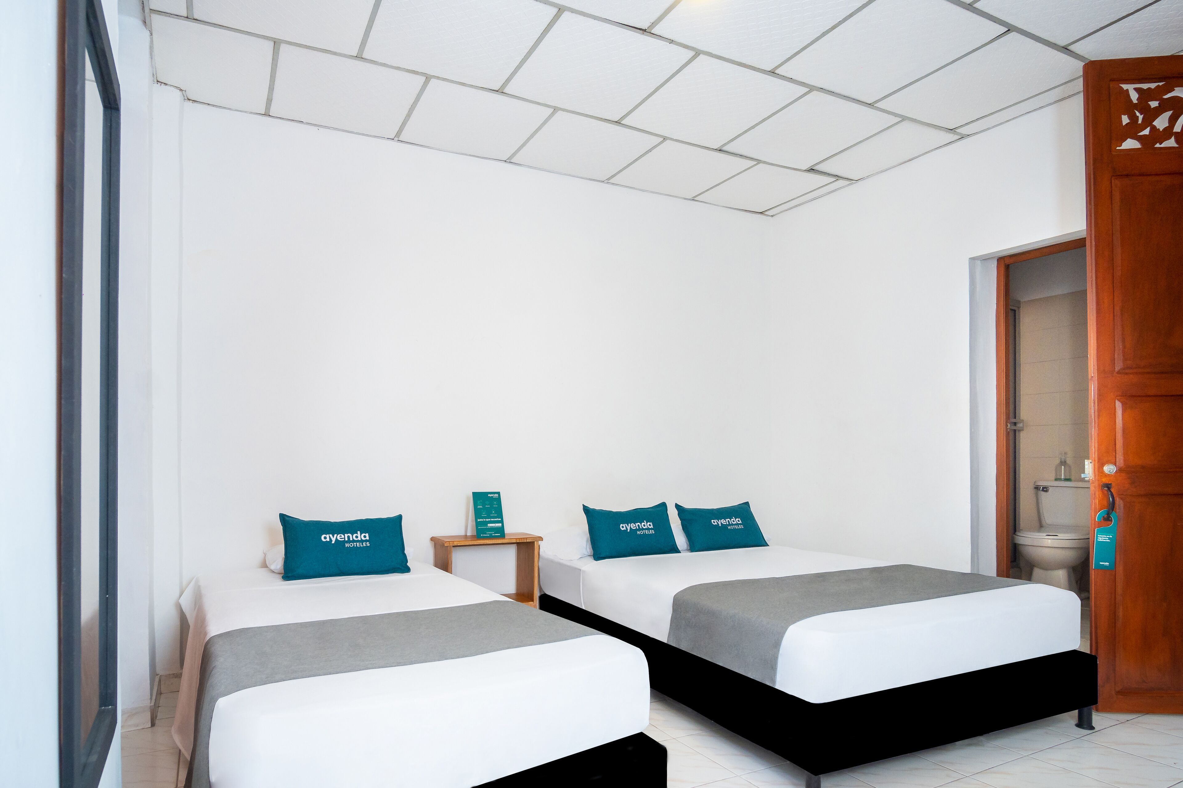 A hotel room with two beds, each with a white mattress and teal pillows, a wooden nightstand, and a wooden door with a handle.