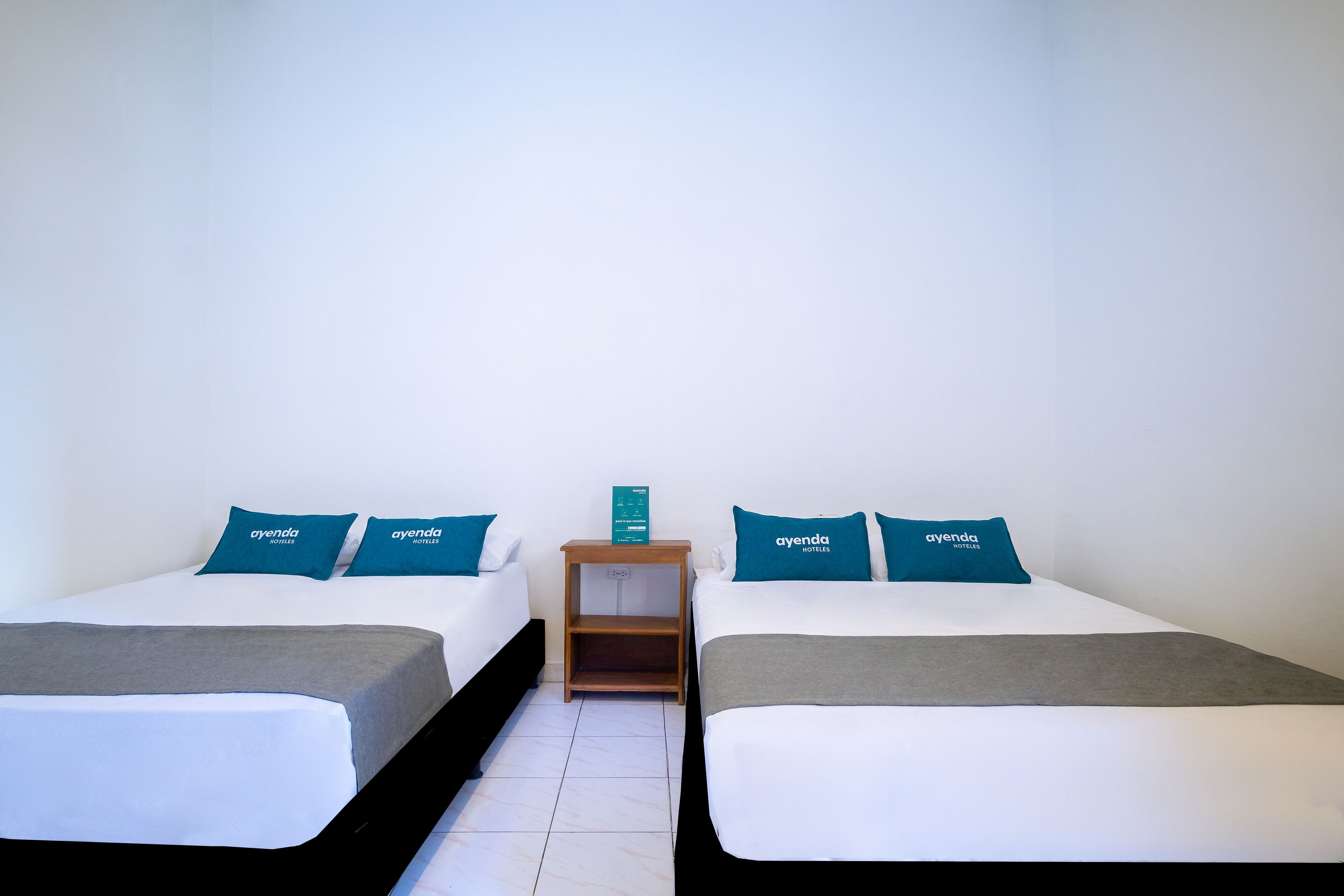 A hotel room with two beds, each with a grey and white bedspread, and teal pillows with the hotel name. There is a wooden nightstand between the beds with a small shelf and a sign.
