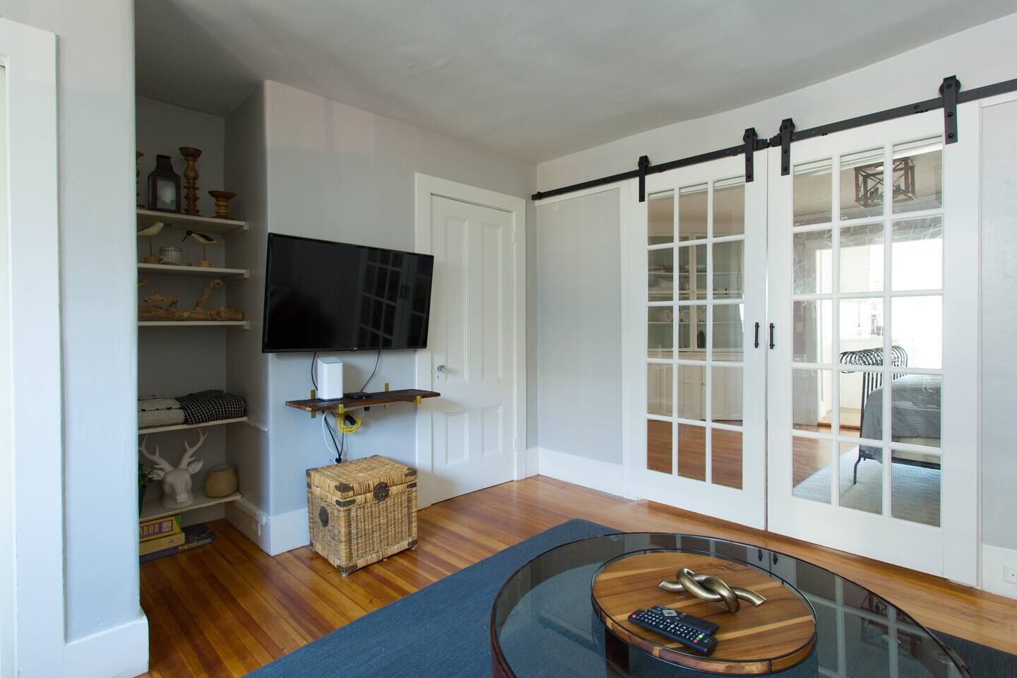 A modern living room with a flat-screen TV mounted on the wall, a glass coffee table, and a sliding glass door leading to a dining area.