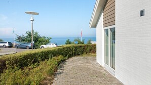Un vialetto lastricato conduce a un edificio con ampie vetrate, con vista sul mare e auto parcheggiate.