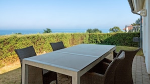 Zona pranzo all'aperto con tavolo e sedie, con vista panoramica sul mare e un siepe.