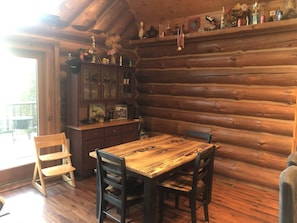 A wooden cabin interior with a dining table and chairs, a wooden cabinet with glass doors, and a shelf with various items.