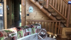 L’intérieur d’une cabane en rondins, avec un escalier en bois, un canapé recouvert d’une couverture à motifs et un panneau d’accueil accroché au mur.