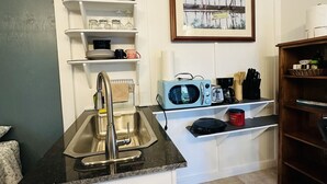 Une cuisine compacte dotée d’un évier en acier inoxydable, de meubles blancs et d’un four à micro-ondes. Un tableau encadré orne le mur, et une petite étagère contient divers ustensiles de cuisine.