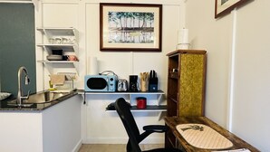 Une cuisine compacte équipée d’un micro-ondes, d’un grille-pain et d’une machine à café sur le plan de travail, d’un fauteuil de bureau noir et d’une petite armoire ornée d’un cadre photo au mur.