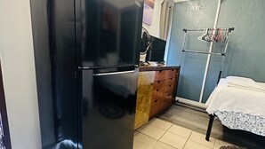 Une cuisine avec un réfrigérateur noir, des placards en bois, un sol carrelé et un lit recouvert d’une couverture blanche.