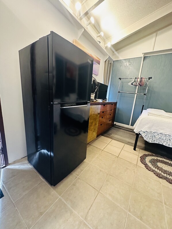A kitchen with a black refrigerator, wooden cabinets, a tiled floor, and a bed with a white cover.