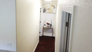 Un couloir étroit aux murs blancs, avec un plancher en bois et une porte sur le côté droit. On aperçoit une petite étagère avec des fleurs au fond.