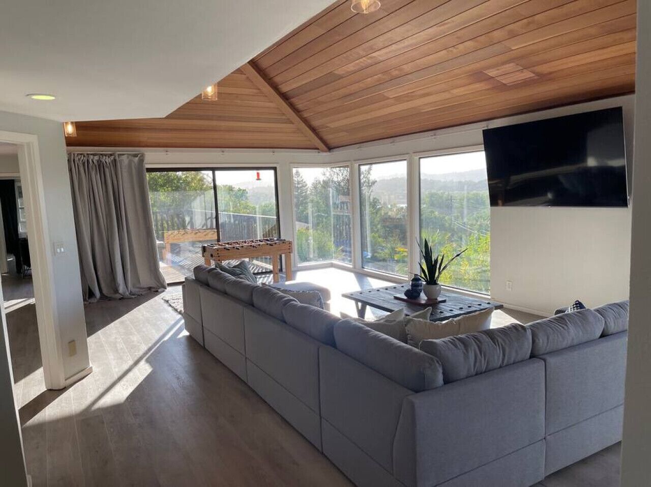 Un salon moderne doté d’un grand canapé d’angle gris, d’un plafond en bois et d’une grande fenêtre donnant sur un paysage verdoyant.
