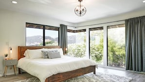 Une chambre avec un grand lit, une table de chevet, une lampe et une vue sur la verdure grâce à de grandes fenêtres.