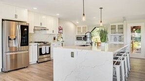 Une cuisine moderne avec des meubles blancs, des appareils en acier inoxydable, un grand îlot central doté de tabourets et un éclairage suspendu.