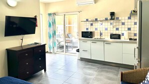 A compact living space with a flat-screen TV, a dark wooden dresser, a wicker chair, a kitchen area with a microwave, toaster, and a refrigerator.