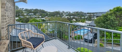 A balcony with wicker furniture, a small round table, and a view of a swimming pool and houses in the background.