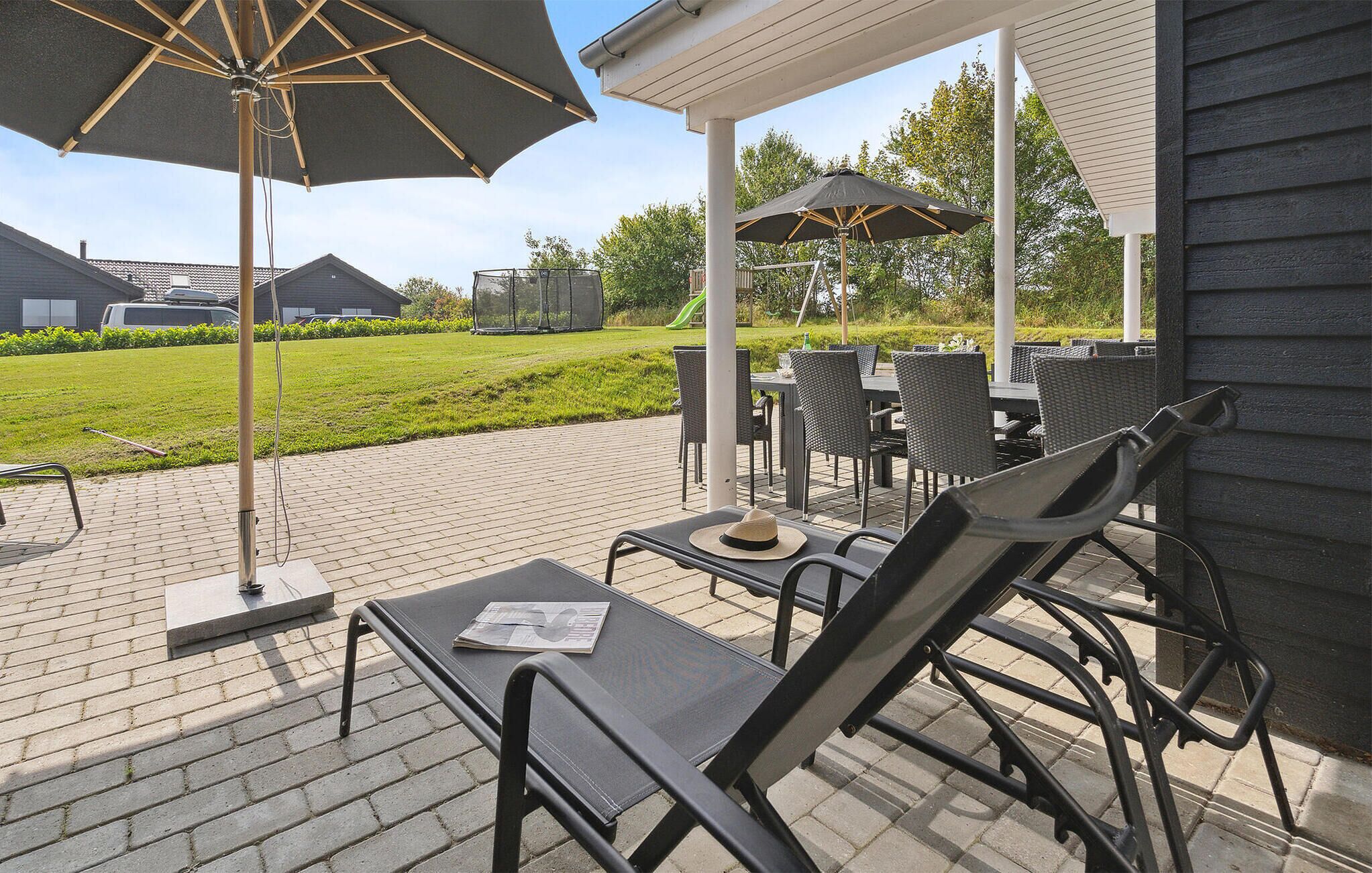 Une terrasse extérieure avec des chaises et des tables en métal noir, un grand parasol et une vue sur une zone verdoyante et des bâtiments en arrière-plan.