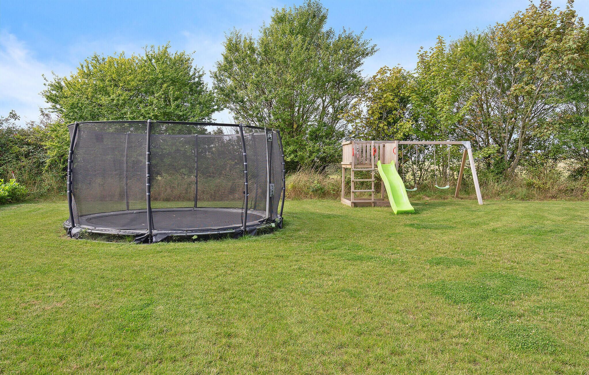 A trampoline with a safety net enclosure and a wooden swing set with a green slide in a grassy yard.
