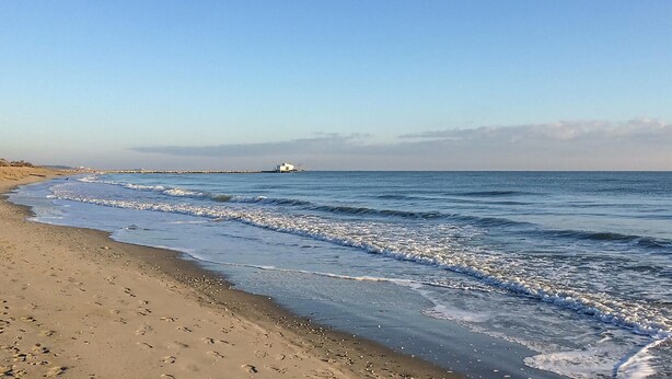 Ein Strand mit einem weißen Gebäude in der Ferne, sandigem Ufer und sanften Wellen.