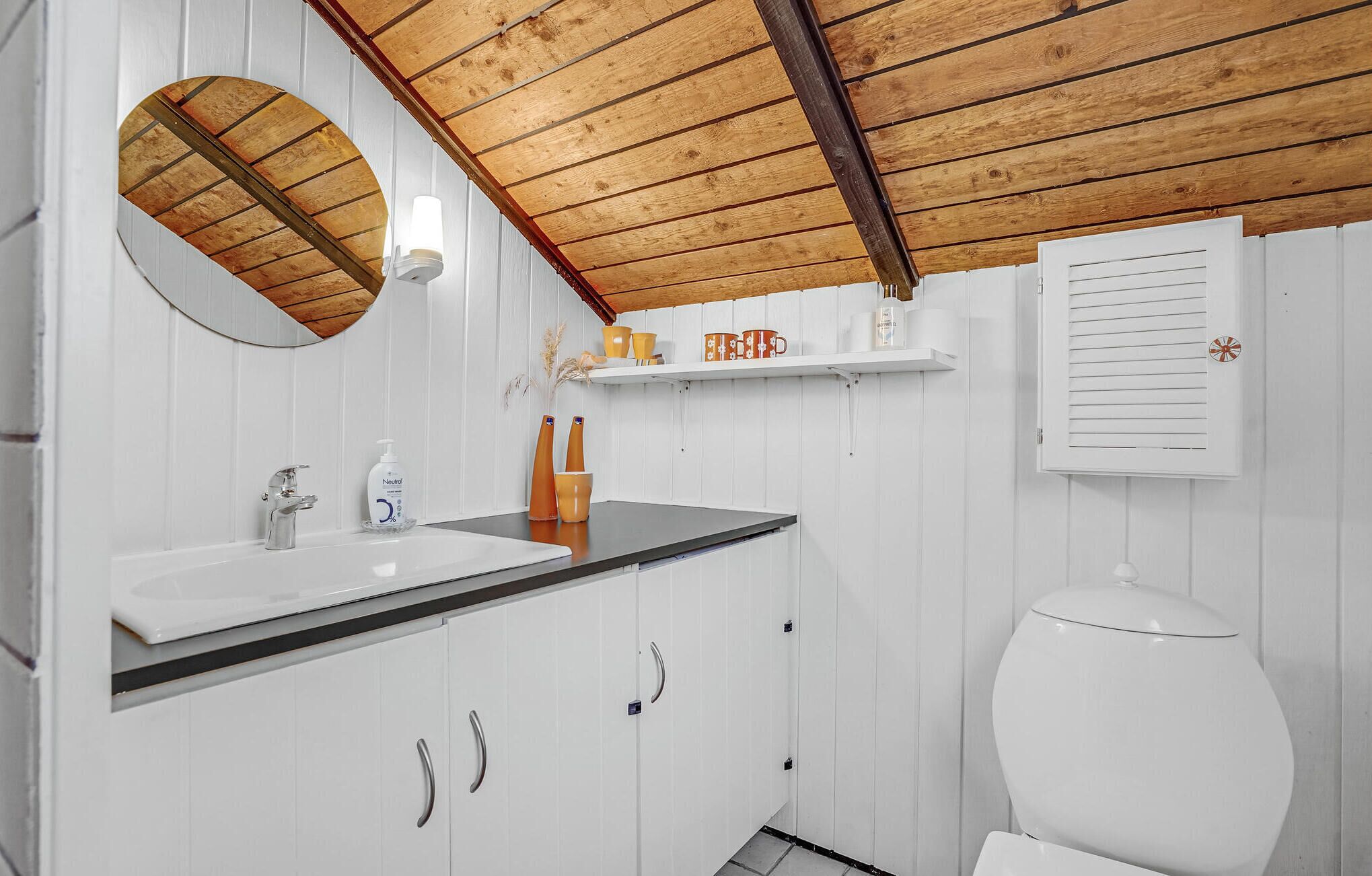 A bathroom with a wooden ceiling, white walls, a round mirror, a sink with a soap dispenser, and a toilet.