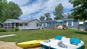 Zona residencial con dos casas, un patio cubierto de césped y un área arenosa con un barco amarillo y uno azul.