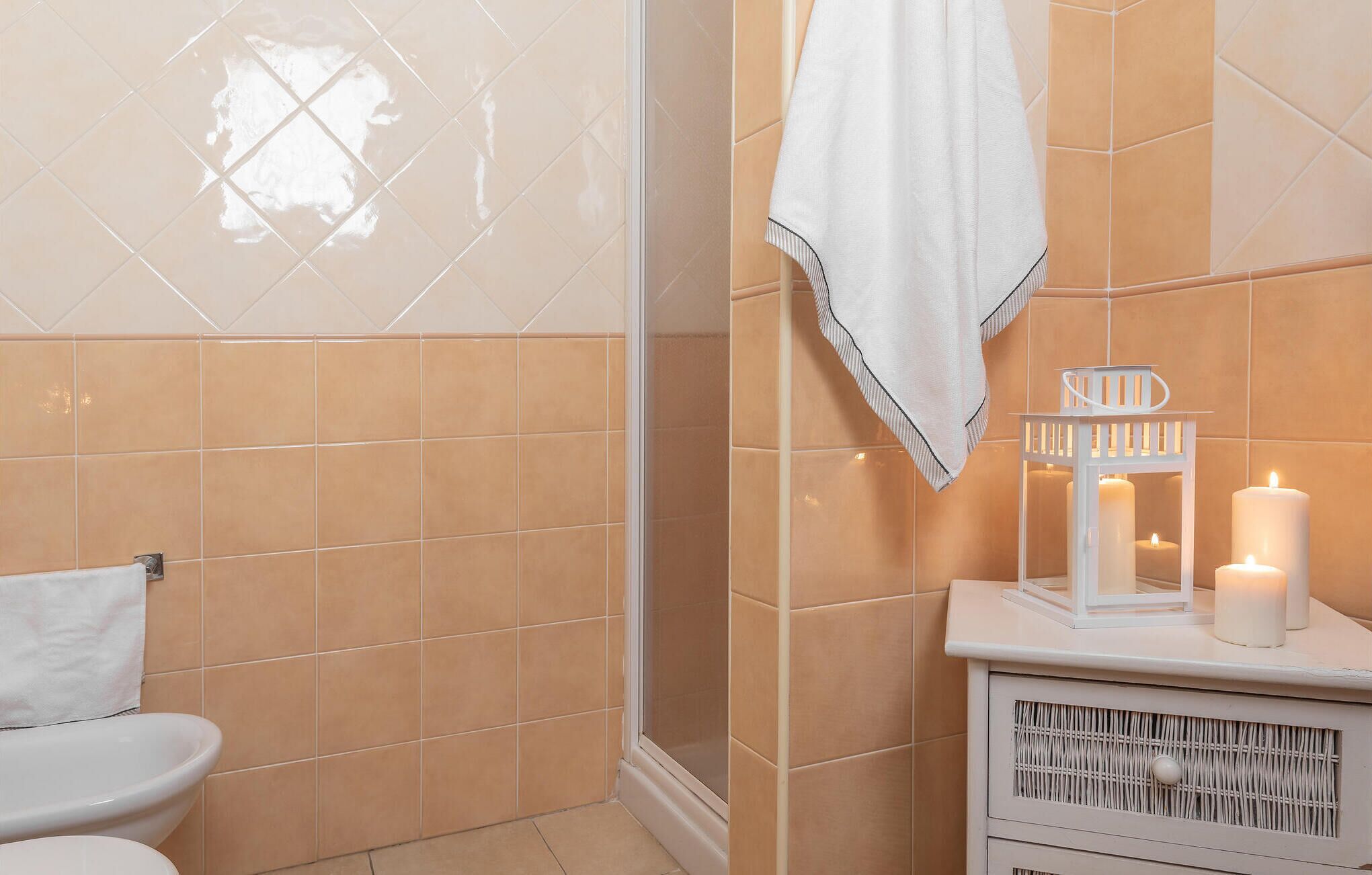 A bathroom with beige tiles, a white toilet, a towel hanging on a rack, and a white cabinet with candles.