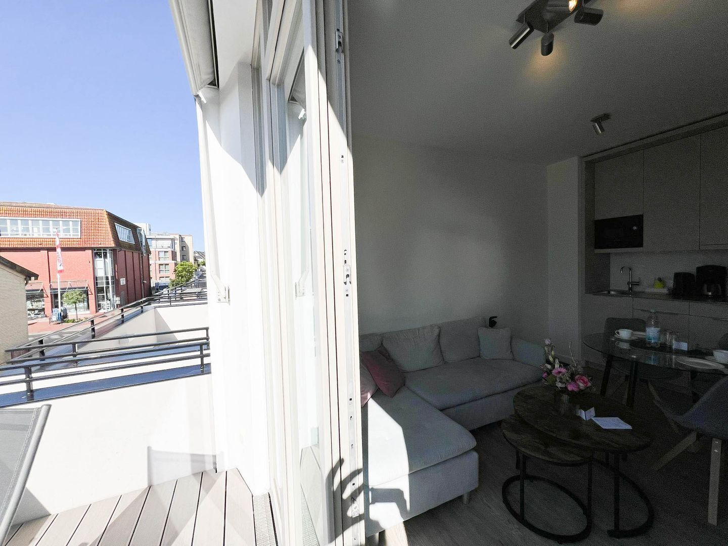A balcony with a white railing, a grey sofa, a small round table with a vase of flowers, and a kitchen area with cabinets and a sink.