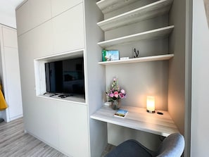 Ein modernes Zimmer mit integriertem Fernseher, einem kleinen Schreibtisch mit Lampe und einem Fenster.