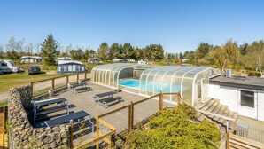 Zona de piscina con una estructura de vidrio, mobiliario exterior y vistas a remolques de campaña al fondo.