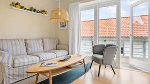 A modern living room with a striped sofa, a wooden coffee table, a grey armchair, and a large window with a view of a red-tiled roof. - 2 bedroom gorgeous home in Gudhjem (Gudhjem)