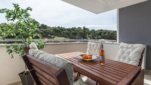 A wooden table with a bowl of oranges and a bottle of wine on a balcony with white cushioned chairs, overlooking a green landscape.