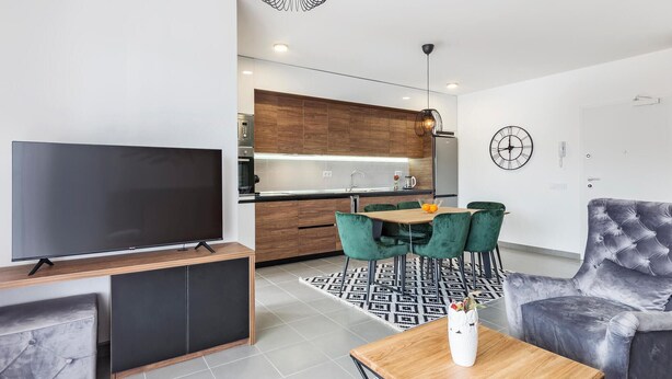 A modern living room with a flat-screen TV, a wooden coffee table, a grey tufted sofa, and a dining area with a table and green chairs.